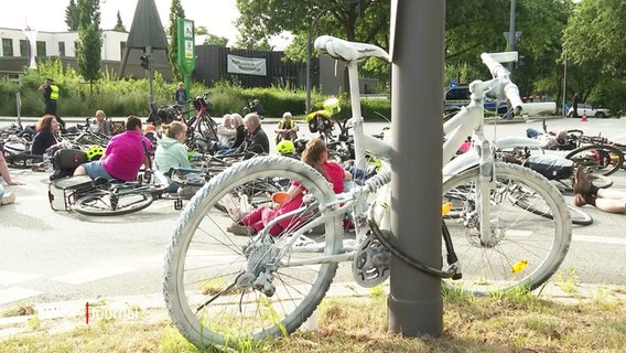 Im Vordergrund das weiß angesprühte Fahrrad der tödlich Verunglückten, im Hintergrund demonstrierende Fahrradfahrer auf der Straße. © Screenshot 