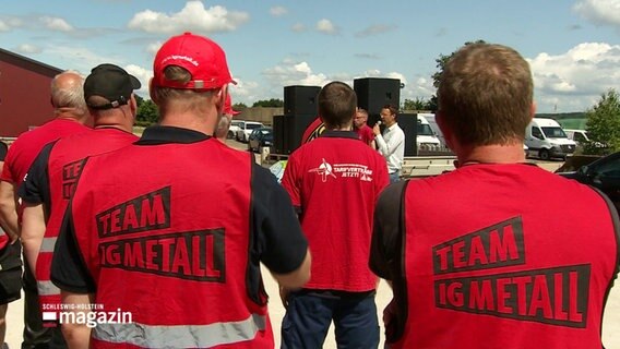 Streikende von hinten in roten Warnwesten mit dem Aufdruck "Team IG Metall" auf dem Rücken. © Screenshot 