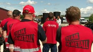 Streikende von hinten in roten Warnwesten mit dem Aufdruck "Team IG Metall" auf dem Rücken. © Screenshot 