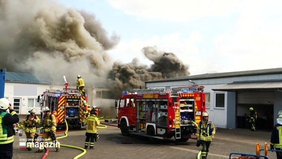 Löschfahrzeuge der Feuerwehr sowie Feuerwehrleute auf einem Gewerbehof, im Hintergrund dicker Qualm und Rauchwolken. © Screenshot 