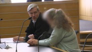 Eine Frau mit unkenntlich gemachtem Gesicht sitzt neben ihrem Verteidiger auf der Anklagebank im Gericht. © Screenshot 