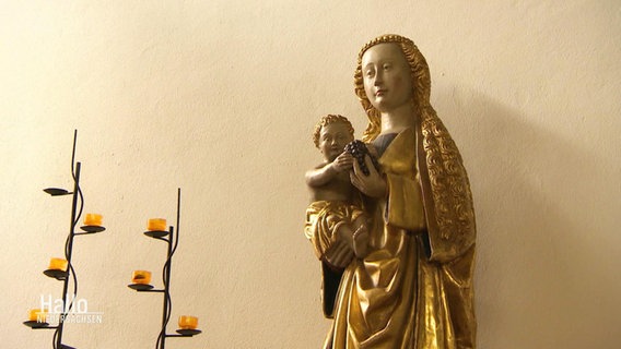 Vor einer weißen Wand steht eine kleinere, vergoldete Statue einer Marifigur mit Jesuskind, daneben zwei moderne Kerzenständer mit Teelichtern. © Screenshot 