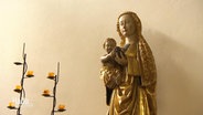 Vor einer weißen Wand steht eine kleinere, vergoldete Statue einer Marifigur mit Jesuskind, daneben zwei moderne Kerzenständer mit Teelichtern. © Screenshot 