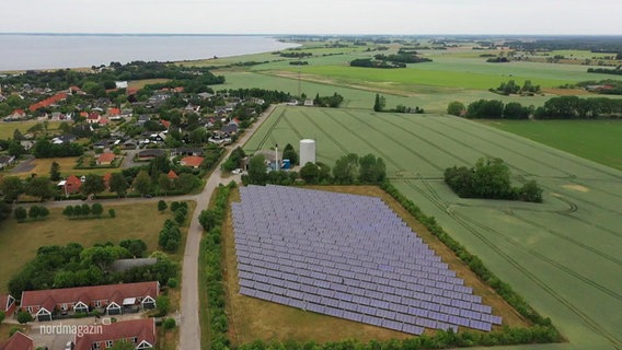 Blick aus der Vogelperspektive: Auf einem Feld steht eine große, mehrreihige Photovoltaikanlage in einer ländlichen Küstengegend. © Screenshot 