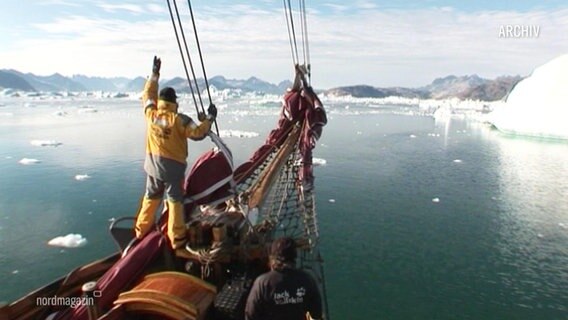 Ein Mann einer Schiffsbesatzung steht in wetterfester Funktionskleidung am Bug eines älteren Schiffes und blickt auf vorausliegende Eisberge im Polarmeer. © Screenshot 