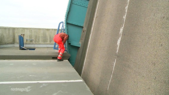 An einer Klappbrücke beugt sich ein Arbeiter an der senkrecht gestellten Fahrbahn untersuchend hinunter. © Screenshot 