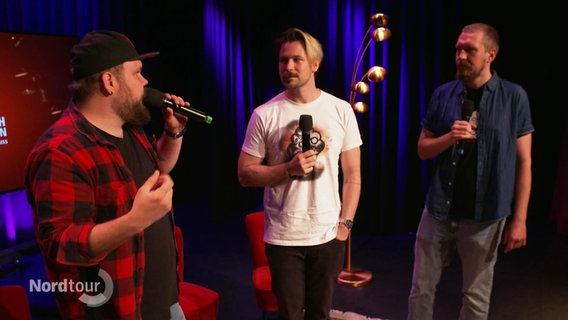 Drei junge Männer mit Mikrofonen stehen auf einer Bühne. © Screenshot 