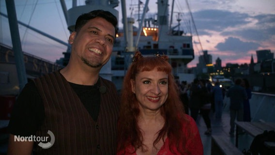 Marcelo Soria und Verónica Villarroel stehen in der Abenddämmerung an Deck der "Cap San Diego". © Screenshot 