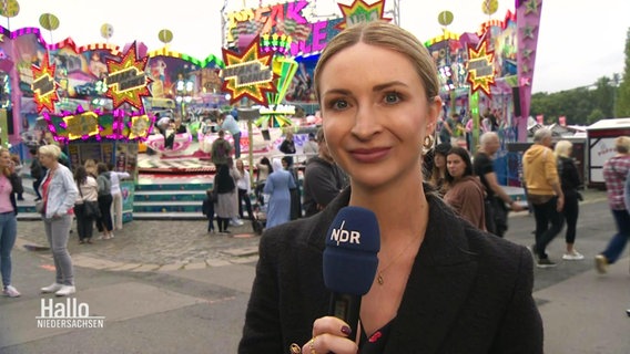 NDR-Reporterin Kerstin Ligendza steht mir Mikrofon vor einem Fahrgeschäft. © Screenshot 