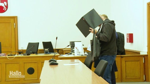 Der Angeklagte verdeckt sein Gesicht hinter einem Aktenordner im Gerichtssaal. © Screenshot 