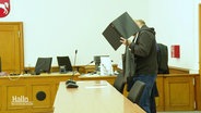 Der Angeklagte verdeckt sein Gesicht hinter einem Aktenordner im Gerichtssaal. © Screenshot 