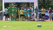 Ein Rugby-Ei liegt auf grünem Rasen. Im Hintergrund laufen zwei Männer-Teams auf den Ball zu. © Screenshot 