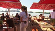 Eine Kamera folgt einer Kellnerin, die sich am Elbstrand zwischen den Stühlen der "Strandperle" hindurchschlängelt. Rote Schirme schützen die Gäste vor der Sonne. Im Hintergrund fließt die Elbe mit Blick auf die Hafenkräne. © Screenshot 