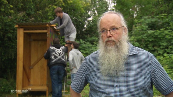 Ein Mann mit einem langen Bart. Im Hintergrund bauen Menschen an einer Hütte. © Screenshot 