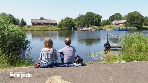 Zwei Personen sitzen am Wasser und genießen das schöne Wetter. © Screenshot 