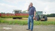Landwirt Eckhard Clausen telefoniert mit einem Handy. Hinter ihm auf dem Feld steht ein Traktor und ein Mähdrescher. © Screenshot 