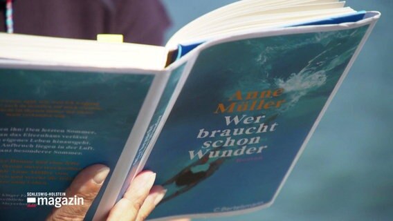 Nahaufnahme von Anne Müllers neuen Roman "Wer braucht schon Wunder". Jemand hält das Buch aufgeklappt zum Lesen in der Hand. Das Cover zeigt eine Unterwasseraufnahme, wie jemand ins Wasser springt. © Screenshot 