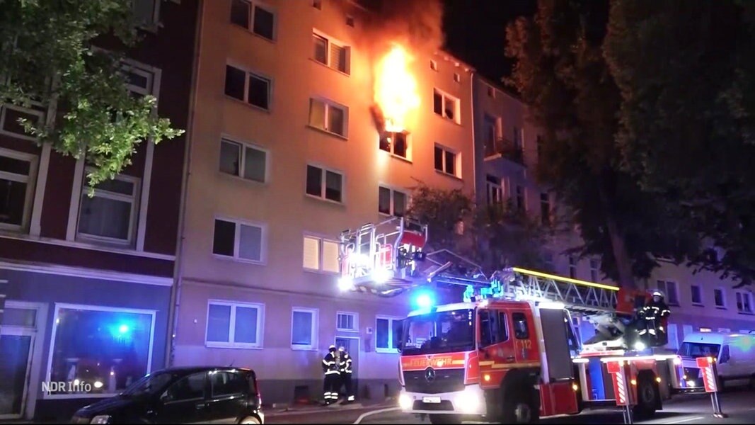Eine Hausfassade, Feuerwehrmenschen und Flammen aus einem Fenster lodernd