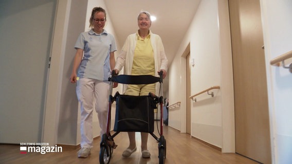 Eine ältere Dame mit Rollator und eine jüngere Pflegerin gehen einen Flur entlang. © Screenshot 