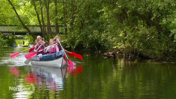 Erwachsene und Kinder paddeln mit einem Kanu durch einen Flusslauf, hinter und rechts von ihnen ist es grün und bewachsen, im HIntergrund eine Fußgängerbrücke. © Screenshot 