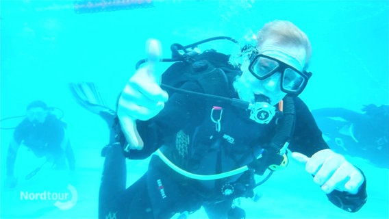 Ein Taucher unter Wasser in einem Schwimmbad. Er schaut direkt in die Kamera und macht ein Handzeichen. © Screenshot 