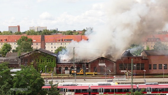 Das Eisenbahnmuseum direkt neben dem Schweriner Hauptbahnhof brennt, das Dach steht in Flammen, dunkler Rauch hängt in der Luft. © Screenshot 