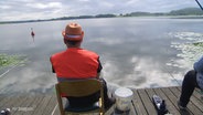 Ein Angler von hinten an einem See. Er sitzt auf einem Stuhl, neben ihm steht ein kleiner Eimer, er trägt einen leuchtend orangen Hut. © Screenshot 