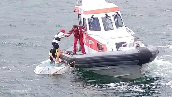 Ein Mann und ein Junge werden von einem kleineren Boot der Seenotrettung von dem Rücken ihres gekenterten Segelboots auf dem Meer gerettet. © Screenshot 