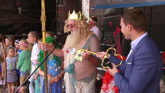 Ein als Neptun verkleideter älterer Mann nimmt auf einer Bühne einen goldenen Schlüssel entgegen. © Screenshot 