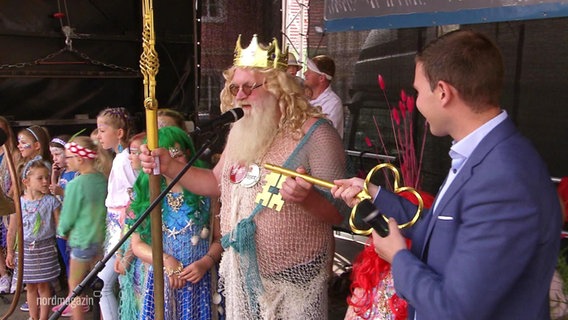 Ein als Neptun verkleideter älterer Mann nimmt auf einer Bühne einen goldenen Schlüssel entgegen. © Screenshot 