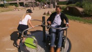 Zwei Menschen spielen an einem Karussell für Rollstuhlfahrer:innen. © Screenshot 