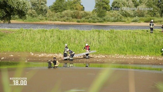 Mehrere Einsatzkräfte der Feuerwehr bergen einen Mann, der hüfttief im Schlick eines kleineren FLussbetts feststeckt. © Screenshot 