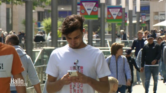 Ein jüngerer Mann schaut beim Gehen in einer Fußgängerzone auf sein Smartphone. © Screenshot 