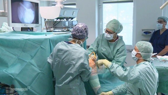 Mehrere Mitglieder eines OP-Teams stehen bei einer Knie-Operation in einem OP-Saal in blauer, steriler Kleidung am OP-Tisch. © Screenshot 