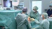 Mehrere Mitglieder eines OP-Teams stehen bei einer Knie-Operation in einem OP-Saal in blauer, steriler Kleidung am OP-Tisch. © Screenshot 
