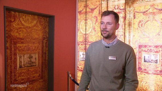 Henning Schröder, Verwaltungsleiter Bernsteinmuseum Ribnitz, im Interview vor einer Bernstein gemusterten Wand © Screenshot 