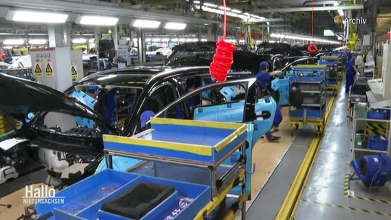Blick in einer Fabrikstraße einer Auto-Fabrik: Mehrere Mitarbeitende werkeln an noch nicht fertiggestellten Automodellen. © Screenshot 