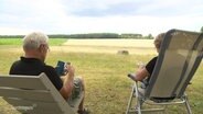 Ein Mann und eine Frau sitzen in Campingstühlen vor einem Kornfeld. © Screenshot 