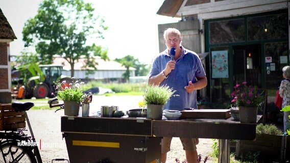 Moderator und Koch Dave Hänsel steht bei sommerlich-sonnigem Wetter vor einem zu einer Außenküche umfunktionierten Lastenfahrrad auf einem dörflichen Platz. © Screenshot 