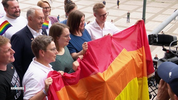 Die zweite Hamburger Bürgermeisterin, Katharina Fegebank, hält mit einigen anderen Personen bei einem Pressetermin eine große Regenbogenflagge auf einem Balkon des Hamburger Rathauses. © Screenshot 