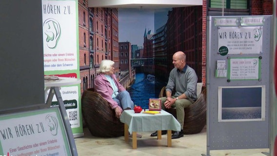 Ein Mann und eine ältere Frau sitzen sich in Sesseln gegenüber in einem Einkaufszentrum, abgeschirmt durch Stelltrennwände. © Screenshot 
