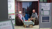 Ein Mann und eine ältere Frau sitzen sich in Sesseln gegenüber in einem Einkaufszentrum, abgeschirmt durch Stelltrennwände. © Screenshot 