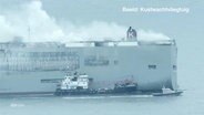 Blick aus der Vogelperspektive auf einen Autfrachter auf offener See von dem hohe Rauchwolken aufsteigen, an seiner Seite hat ein kleineres Schiff festgemacht. © Screenshot 