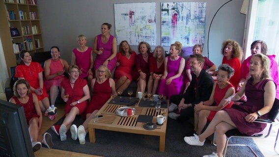 Choristinnen des Damen-Likör-Chors, alle in Pink- und Rottönen gekleidet, in einem Wohnzimmer auf der Couch. © Screenshot 