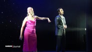 Ein Mann und eine Frau auf einer Bühne- Sie trägt ein pinkes Kleid und streckt ihm ihre linke Hand entgegen, singt aber in die entgegengesetzte Richtung. Er trägt einen Anzug und schaut geradeaus. © Screenshot 