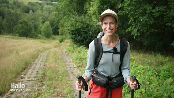 Katharina Trabert lächelt in die Kamera, sie hat eine Baseballkappe auf, Rucksack und Wanderstöcke dabei. © Screenshot 