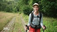 Katharina Trabert lächelt in die Kamera, sie hat eine Baseballkappe auf, Rucksack und Wanderstöcke dabei. © Screenshot 