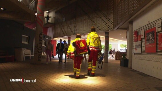 Von hinten gesehen: Zwei junge Menschen in der Arbeitskleidung von Rettungssanitätern gehen durch ein Gebäude. © Screenshot 