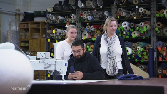 In einer Polster-Werkstatt: Die Auszubildende Ann-Charlott Moll mit ihrer Chefin. Im Vordergrund sitzt ein Mann an einer Nähmaschine. © Screenshot 