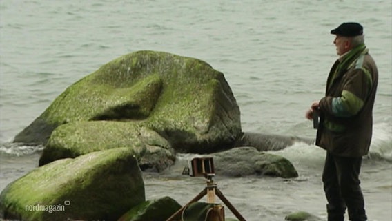 Der Fotograf Volkmar Herre steht am Ufer der Ostsee neben großen Findlingen und schaut aufs Wasser, links neben ihm seine Camera Obscura. © Screenshot 